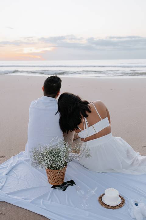 Foto do casal sentado de costas na areia observando o mar.'