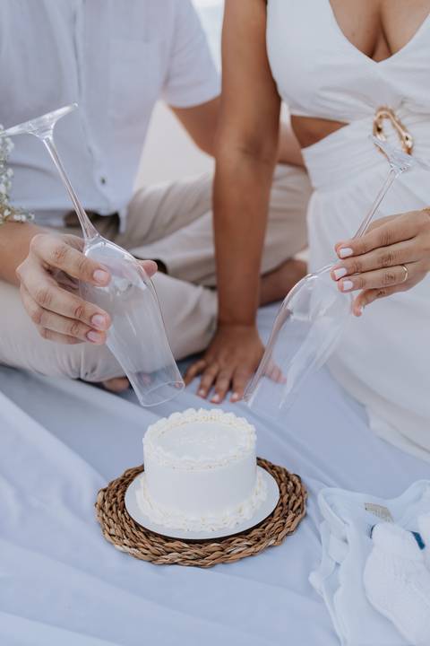 Foto de detalhe das mãos do casal posicionando as taças para o corte do bolo.'