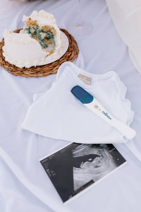 Foto em detalhe da roupinha branca e o teste de gravidez ao lado do ultrassom.'