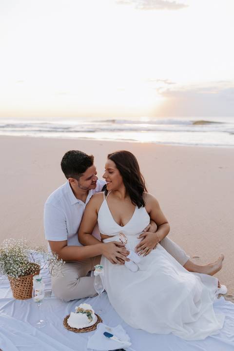 Foto do casal abraçado na areia celebrando a novidade no nascer do sol.'