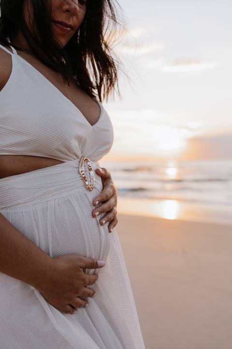 Foto aproximada da mão da gestante sobre a barriga com a luz dourada ao fundo.'