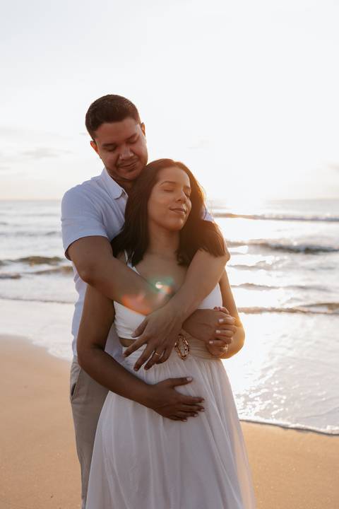 Foto do pai abraçando a gestante por trás com os olhos fechados.'