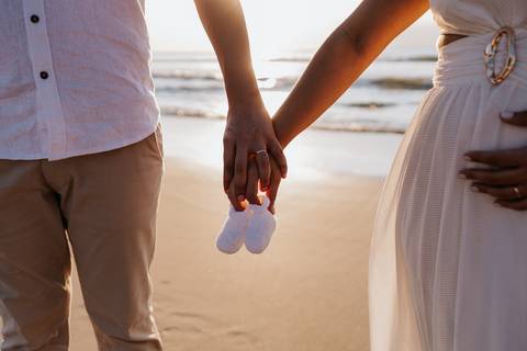 Foto em detalhe das mãos do casal segurando sapatinhos de bebê na areia.'