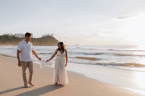 Foto do casal caminhando de mãos dadas segurando os sapatinhos.'