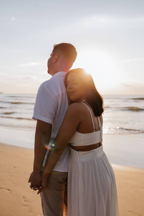 Foto da gestante abraçando o parceiro por trás sob a luz dourada.'
