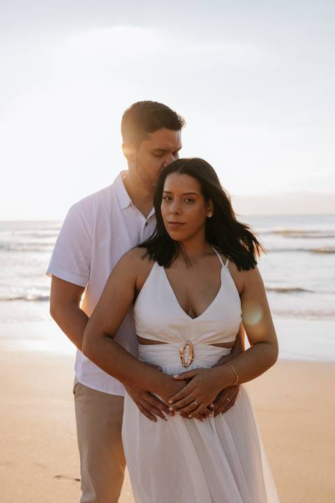 Foto do casal posando na beira do mar durante o nascer do sol.'