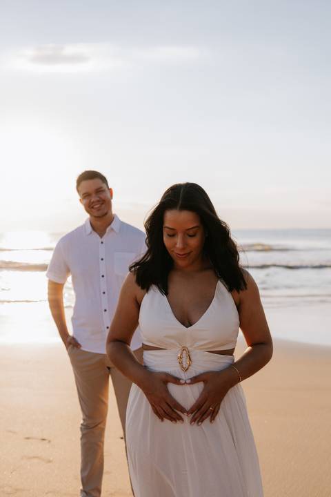 Foto da gestante olhando para a barriga com o parceiro ao fundo.'
