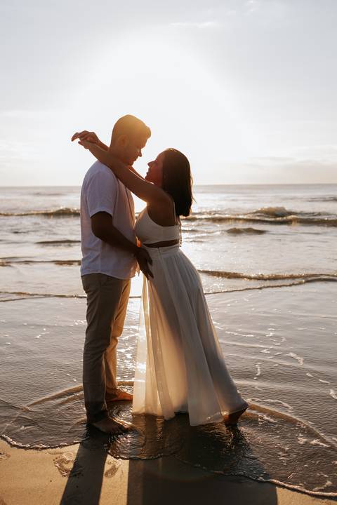 Foto do casal abraçado na areia com o mar ao fundo.'