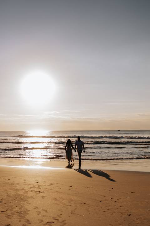 Foto da silhueta do casal caminhando na praia sob o sol intenso.'
