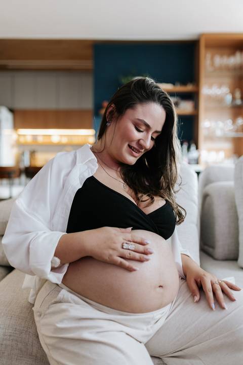 Foto da gestante sentada no sofá olhando para a barriga em ensaio em casa.'