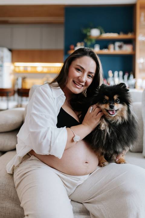 Foto da gestante e do cão sorrindo para a câmara no sofá.'
