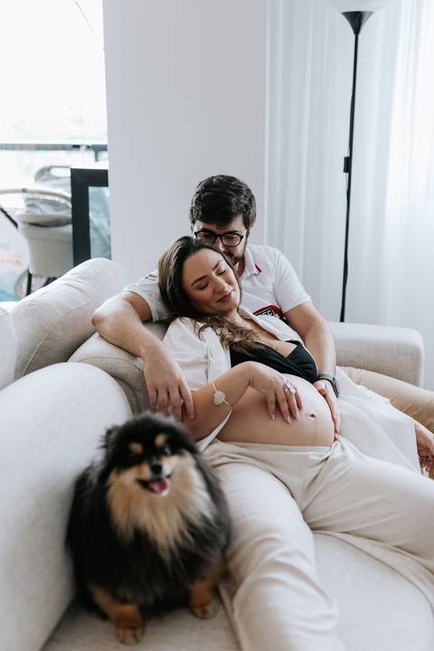 Foto do casal abraçado no sofá com a barriga de grávida em destaque.'