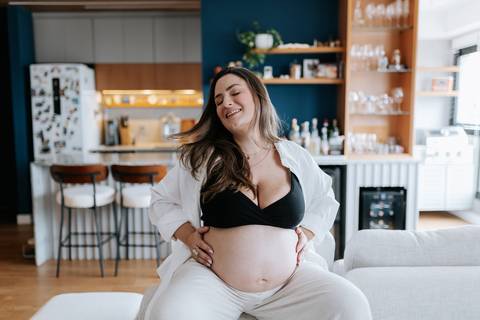 Foto da gestante sentada no sofá com as mãos na barriga e camisa aberta.'