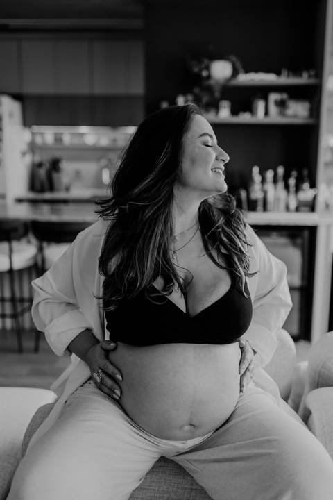 Foto em preto e branco da gestante sentada no sofá destacando a barriga.'