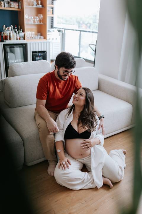 Foto do casal sorrindo e interagindo sentado no chão ao lado do sofá.'
