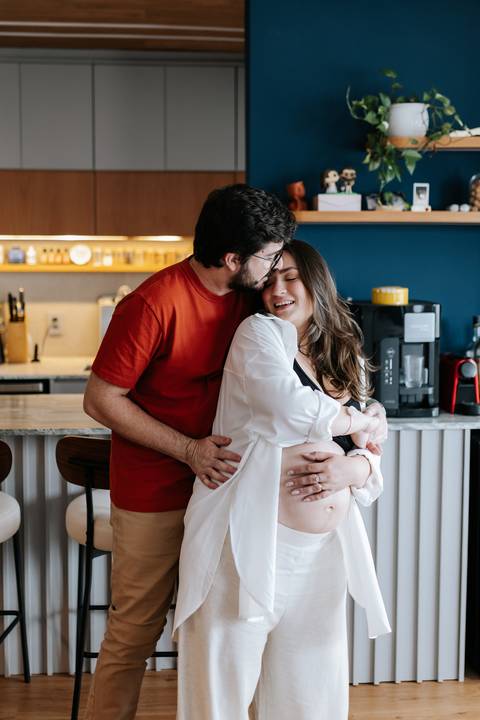 Foto do casal abraçado na cozinha durante ensaio gestante em casa.'