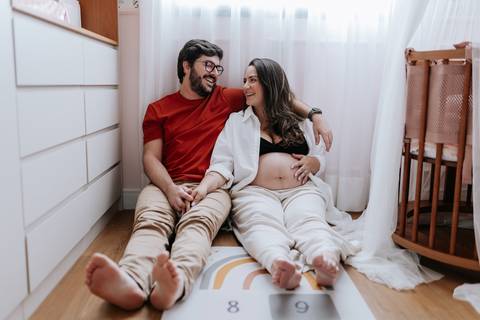 Foto do casal sentado no chão do quarto do bebê com a barriga à mostra.'