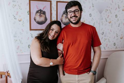 Foto do casal sorridente posando no quarto decorado para a chegada da filha.'