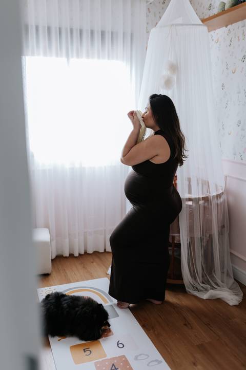 Foto da silhueta da gestante em frente à janela beijando a roupinha do bebê'