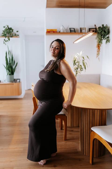 Foto da gestante encostada na mesa de jantar realçando a barriga de grávida.'