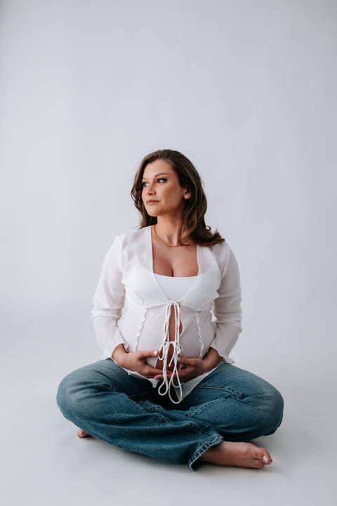 Foto de perfil da gestante com blusa branca destacando a barriga em fundo neutro.'