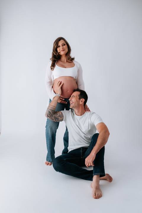 Foto do pai sentado abraçando a barriga da gestante em pé no estúdio.'