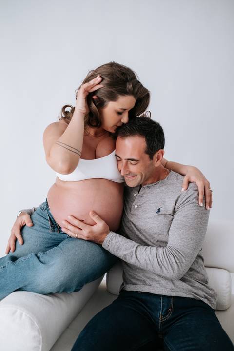 Foto do casal sentado no chão abraçando a barriga em momento afetivo.'