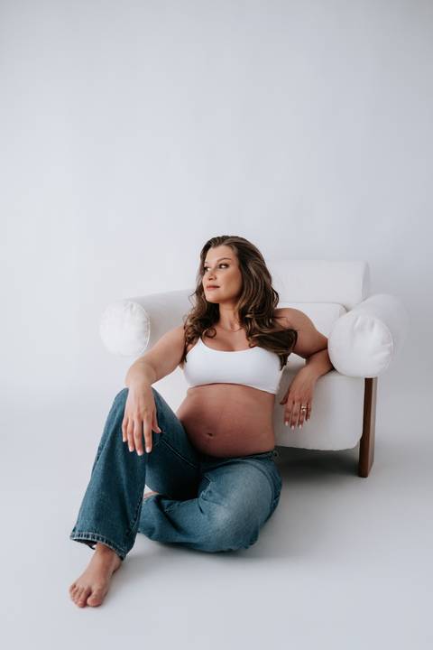 Foto da gestante sentada no sofá branco com look casual de top e calça jeans.'