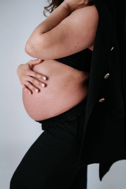 Foto em detalhe da mão da gestante sobre a barriga com blazer preto.'