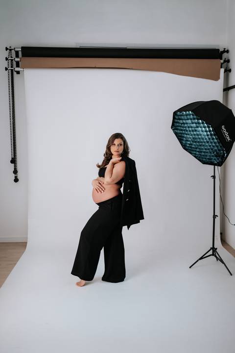 Foto da gestante de perfil com blazer preto nos ombros e luz de estúdio ao fundo.'