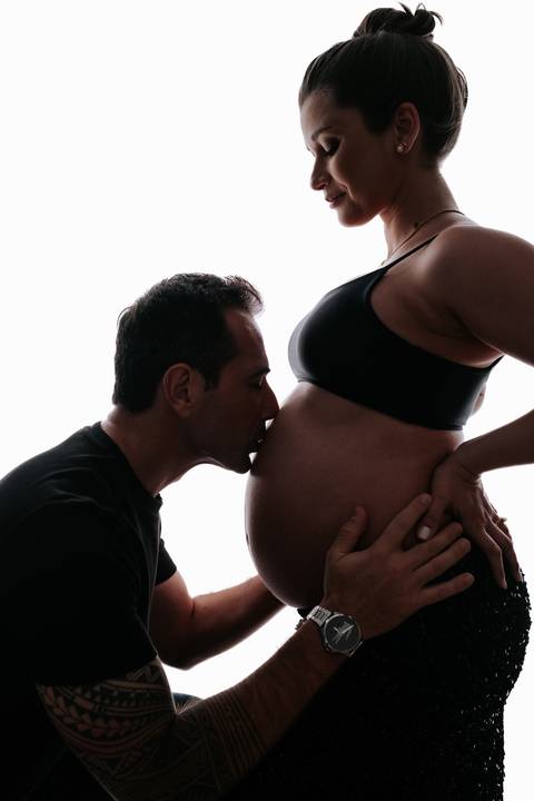 Foto da silhueta do pai beijando a barriga da gestante contra um fundo branco iluminado.'