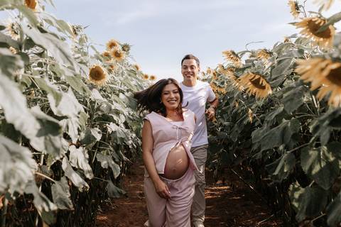 Foto da gestante sorrindo com o parceiro ao fundo em meio aos girassóis.'