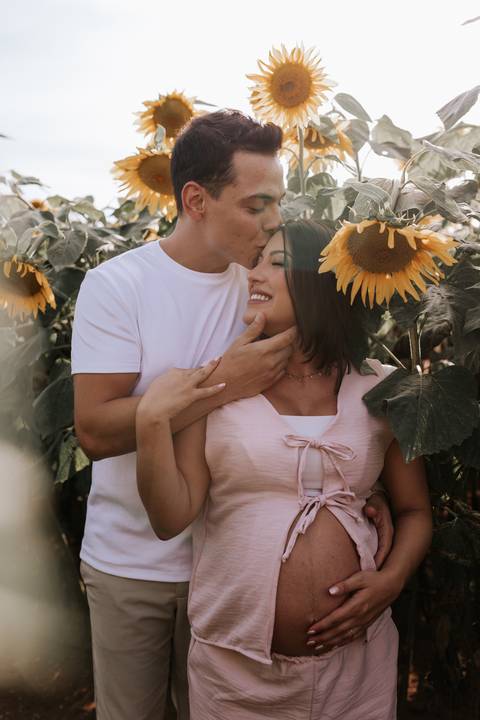 Foto do pai beijando a testa da gestante cercados por girassóis.'