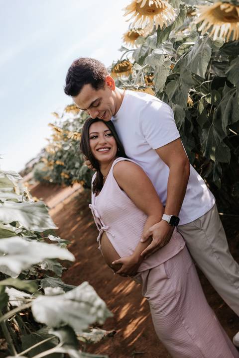 Foto da gestante no campo com o parceiro a abraçando por trás.'