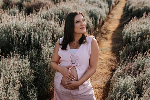 Foto da gestante com as mãos na barriga apreciando o cenário de flores roxas.'