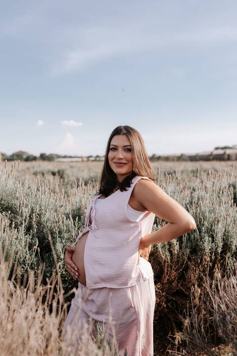 Foto da gestante sorridente no campo de lavandas com as mãos na cintura.'