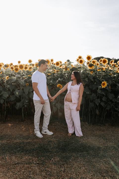Foto do casal de mãos dadas em frente ao campo de girassóis em luz natural.'