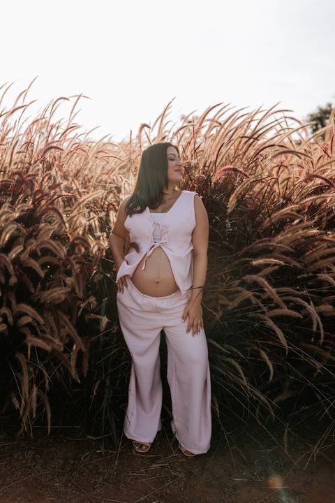 Foto da gestante em pé entre capins dourados destacando a barriga de grávida.'