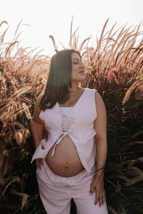 Foto aproximada da gestante sorrindo em meio à vegetação seca no entardecer.'