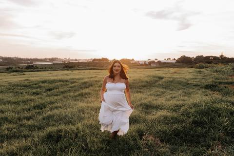 Foto da gestante caminhando em um campo aberto com vestido branco fluido.'