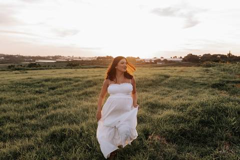 Foto da gestante sorrindo enquanto caminha no gramado durante o pôr do sol.'