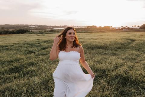 Foto aproximada da gestante com vestido branco e mãos na barriga sob a luz dourada.'