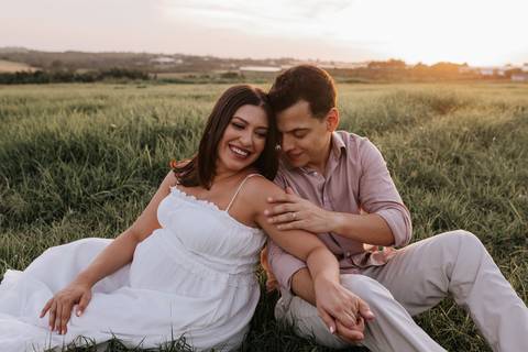 Foto do casal sorrindo e relaxado sentado no gramado em ensaio externo.'
