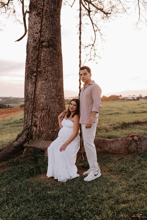 oto da gestante sentada sob uma árvore com o parceiro ao lado.'