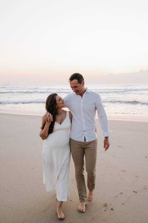 Foto do casal caminhando abraçado na beira do mar durante o nascer do sol.'