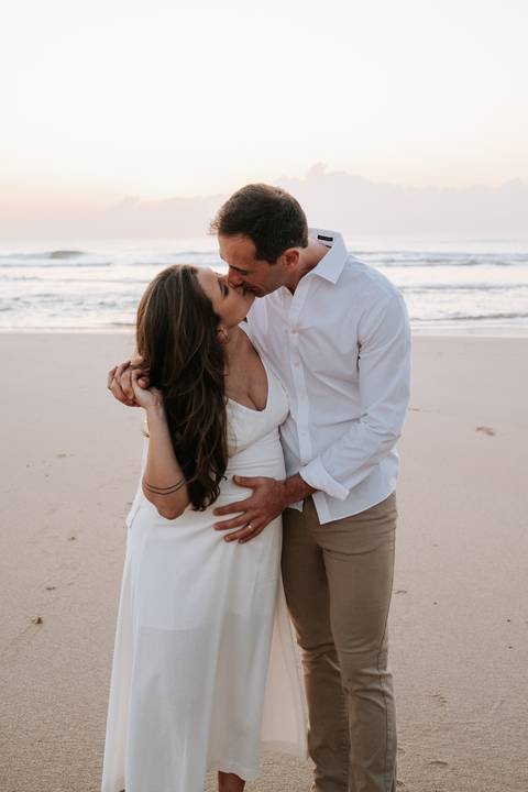 Foto do casal se beijando com o mar e o céu claro ao fundo.'