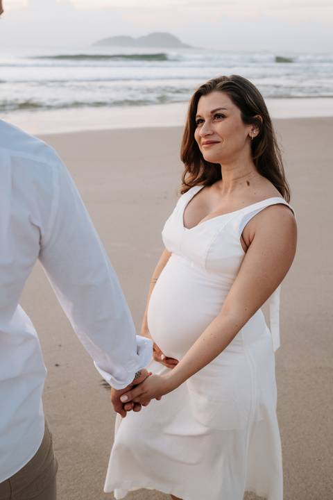 Foto de detalhe da barriga da gestante com vestido branco e o parceiro ao lado.'