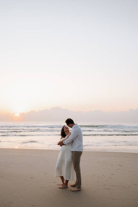 Foto em plano aberto do casal abraçado na areia com o sol surgindo no horizonte.'