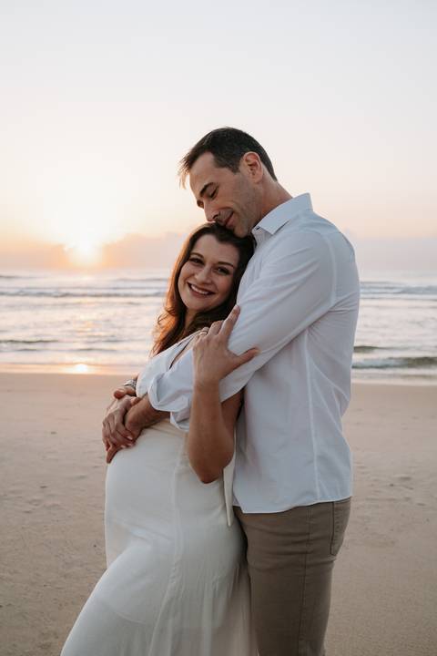 Foto da gestante sorrindo enquanto é abraçada por trás pelo parceiro sob a luz dourada.'
