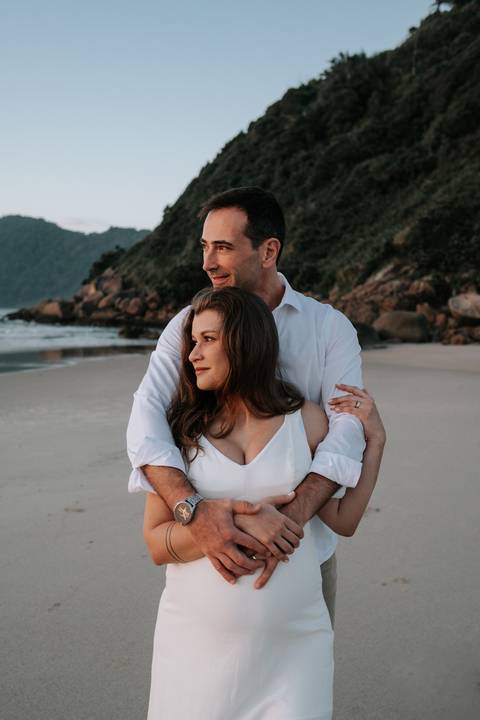 Foto do pai abraçando a gestante por trás com as rochas e o mar ao fundo.'